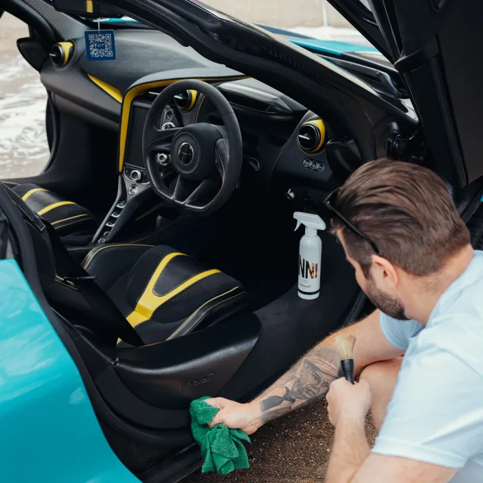 A person cleans a sports car's interior with a brush and cloth. The car has black and yellow seats, and a spray bottle labeled "NNH!" rests on the floor mat.