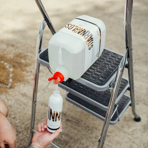 A large bottle labeled "SILIKANGLOS" dispenses liquid into a smaller bottle labeled "INN" via a red nozzle, placed on a step ladder, outdoors.