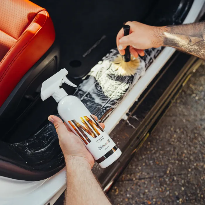 A person holds a white spray bottle labeled "The Last Coat" while applying foam cleaner with a brush to a car's interior door frame near a red seat.