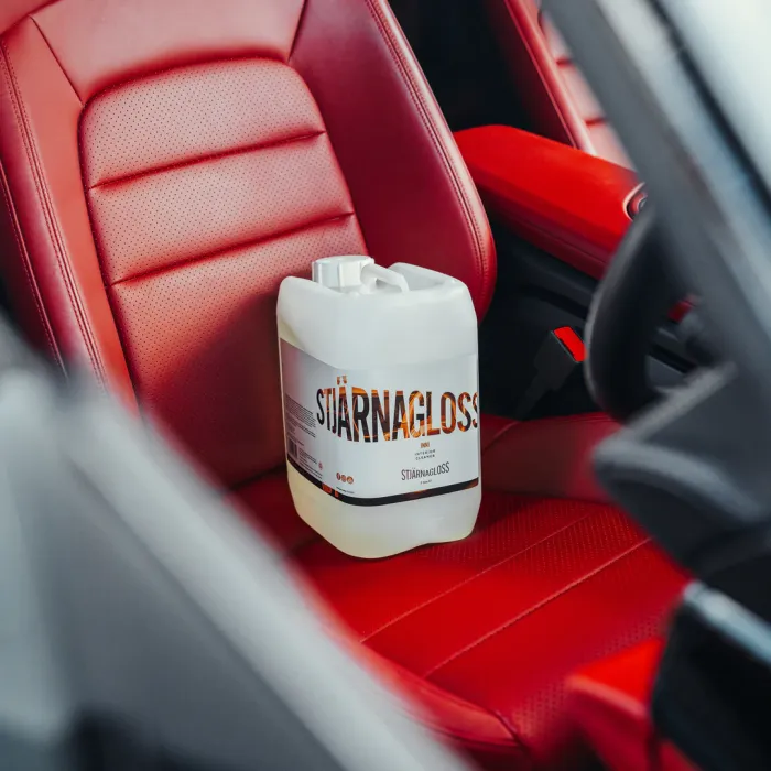 A plastic container labeled "Stjärnagloss" rests on a red leather car seat, set inside a vehicle's interior.