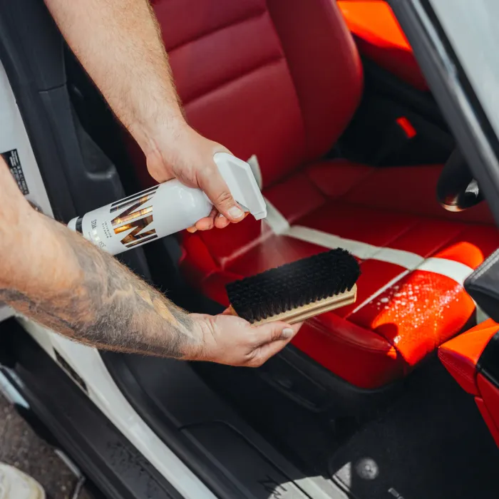 Tattooed arm sprays cleaning solution onto a black bristle brush while standing next to a car with red leather seats.