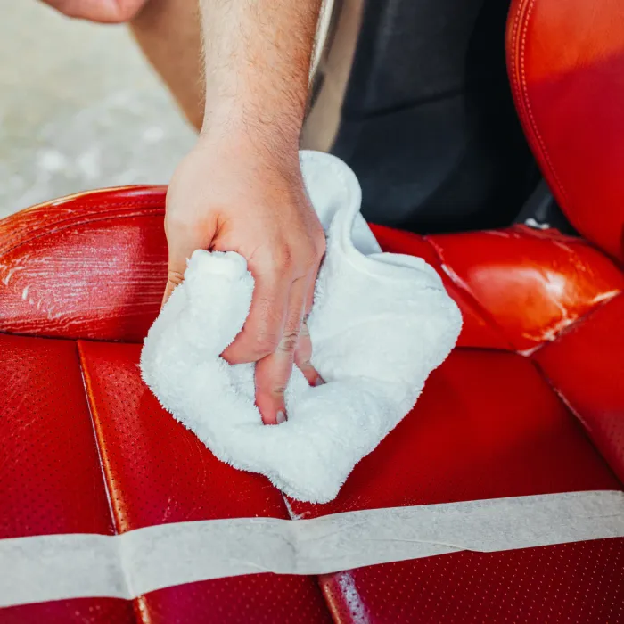 A hand holding a white cloth wipes the surface of a red leather car seat, with some cleaning product applied, inside a vehicle.