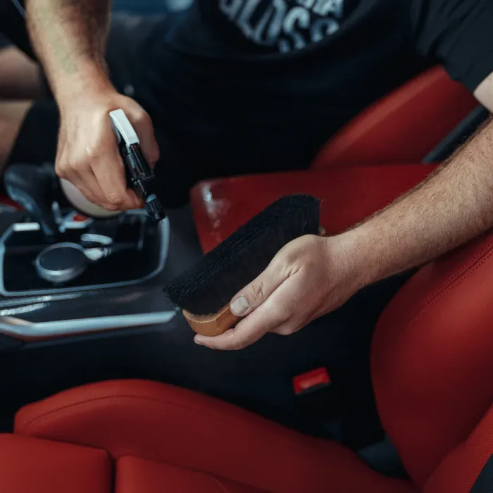 A hand sprays a cleaning solution onto a black-bristled brush inside a car with red leather seats and other visible car controls in the background.