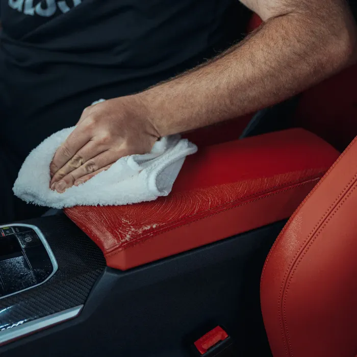 A hand wipes a red leather car armrest with a white cloth, inside a vehicle with a black interior and visible central console buttons.