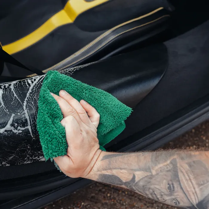 A hand uses a green cloth to clean a black car seat inside a vehicle, with visible tattoos on the arm.