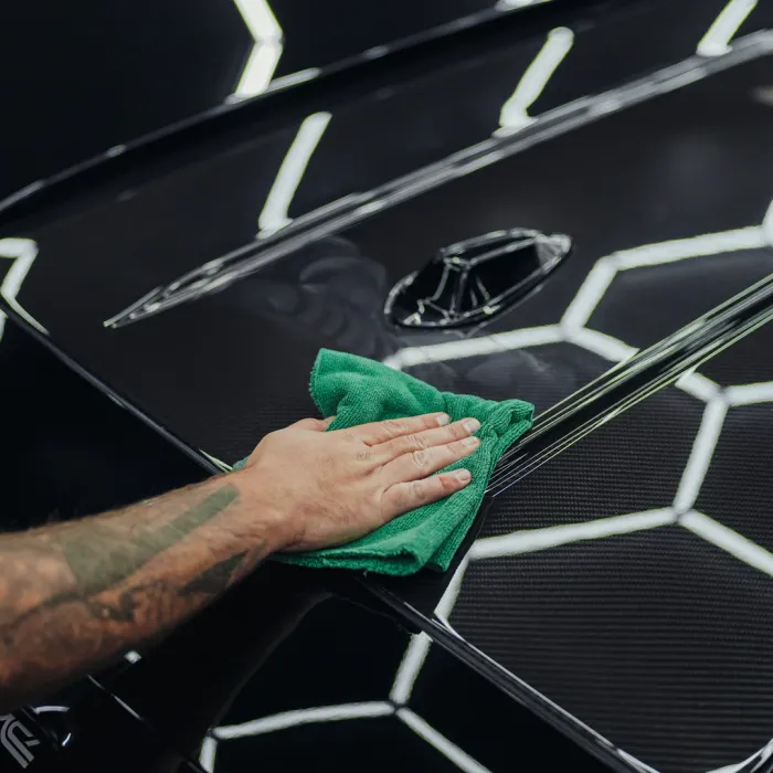 A tattooed hand cleans a car's glossy, black surface with a green cloth in a well-lit area featuring geometric lighting reflections.