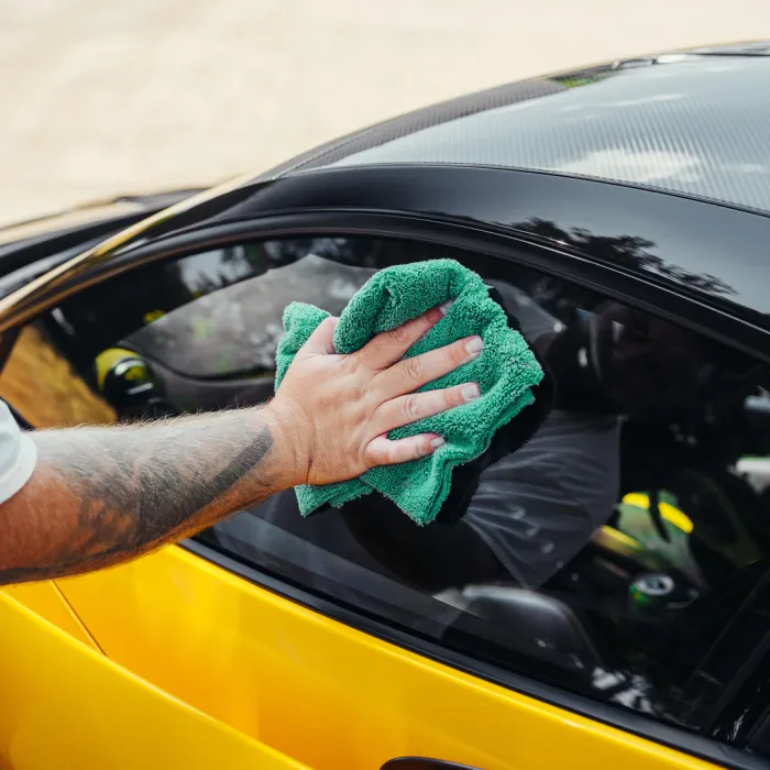 A tattooed hand is cleaning the window of a yellow car with a green microfiber cloth outside on a sunny day.