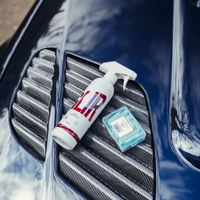 A bottle of "CLAY LUBRICANT" and a blue plastic container rest on a shiny, vented car hood in an outdoor setting.