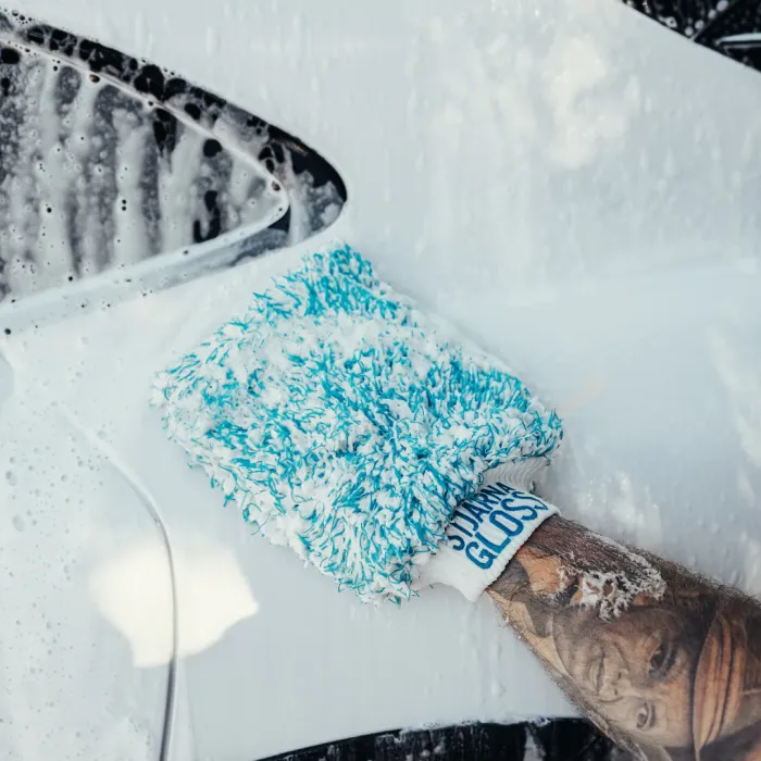 A tattooed arm uses a soapy microfiber glove labeled "SUDS GLOVE" to wash a white car, with the vehicle surface covered in foam.
