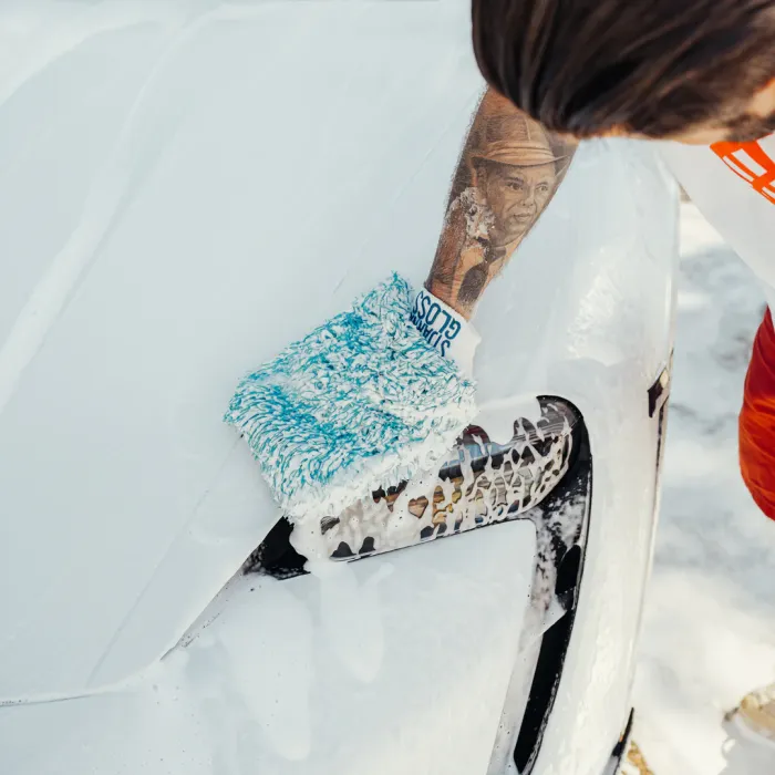 A gloved hand with a tattoo washes a foamy white car. The glove reads "SUDS GLOSS." The car is outdoors, and only the front section is visible.