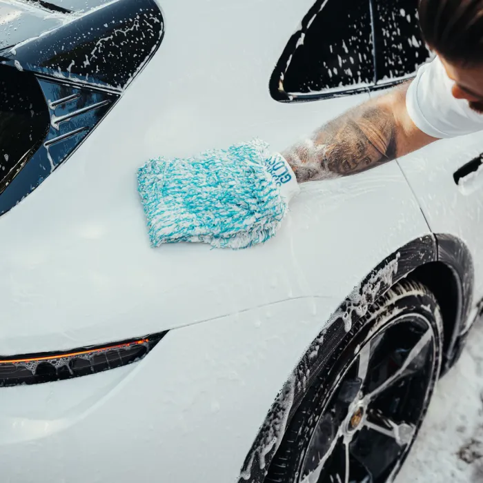A person’s soapy gloved hand scrubs a sleek white car with black accents, featuring suds on the vehicle's surface in an outdoor environment.