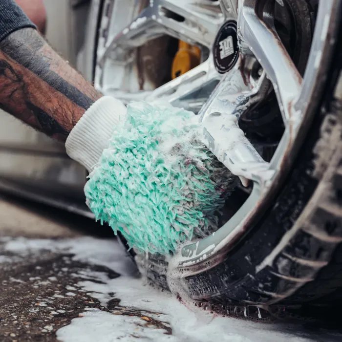 Arm cleans a car wheel using a soapy green brush mitt; the tire and chrome wheel are partially covered in foam, situated on a wet, soapy garage floor.