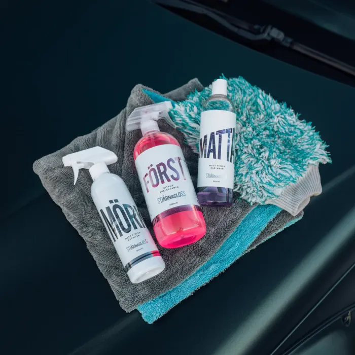 Three car cleaning products labeled "MÖRK," "FÖRST," and "MATTI" are placed on top of gray and turquoise cleaning cloths, resting on a dark car surface.