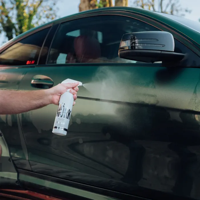 A hand sprays cleaning solution from a bottle labeled "SNEAK" onto the side of a dark green car outdoors, with trees visible in the background.