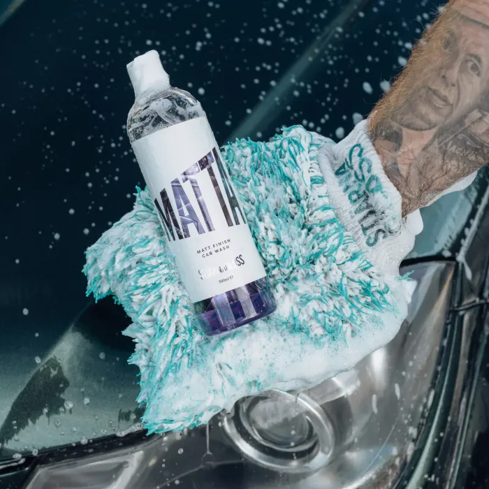A bottle of car wash labeled "MATT" rests on a sudsy cleaning mitt worn by a tattooed arm near a wet car surface. The label reads "MATT FINISH CAR WASH" and "500 ml / 16 fl oz."