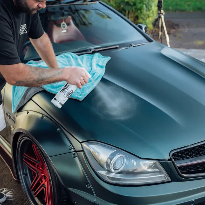 A person sprays and wipes the hood of a dark green car with a light blue microfiber cloth outdoors, with greenery and equipment in the background.