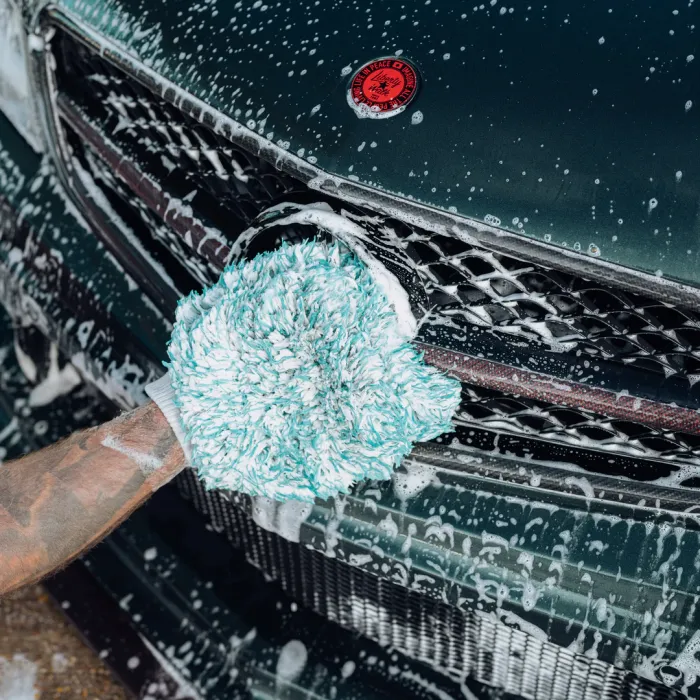 A gloved hand scrubs a soapy car grille with a green and white sponge. A red emblem with the text "Let the peace of adventure" decorates the car's hood.