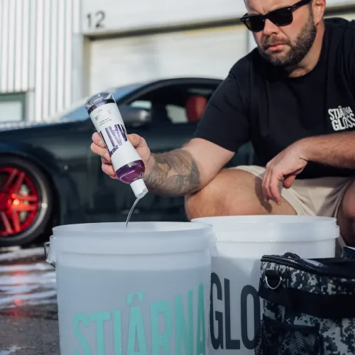A man wearing sunglasses pours Stjärnagloss shampoo into a large white bucket. He is kneeling next to a black car with red wheels in an outdoor setting.