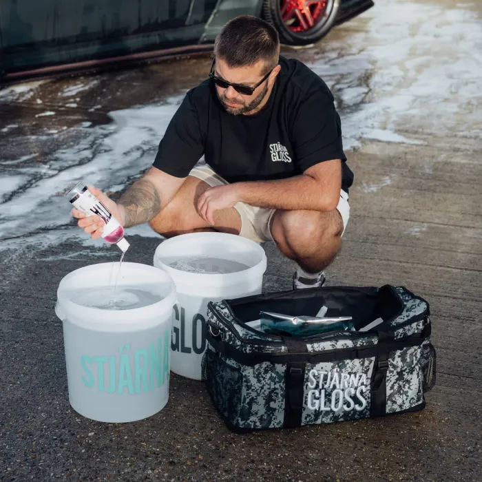 A man pours car wash solution into one of two white buckets labeled "STJÄRNAGLOSS" on a wet pavement beside a parked, partially visible car and a branded duffel bag.
