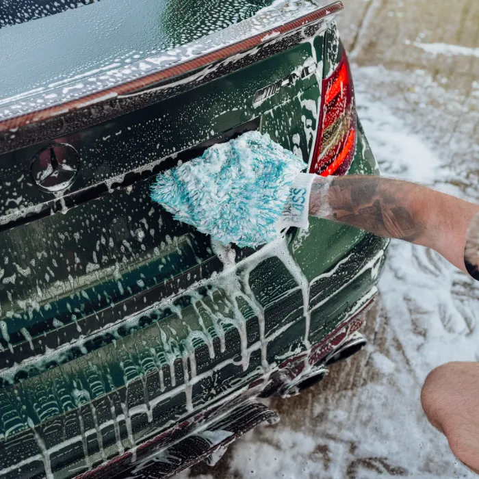 A person wearing a soapy wash mitt cleans the rear end of a dark green Mercedes car, covered in suds, with a concrete pavement in the background.