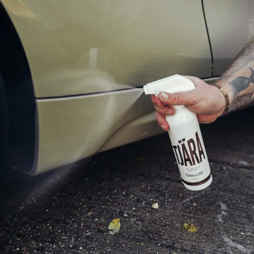 A hand sprays cleaning solution from a bottle labeled "TJÄRA" onto a car's lower side panel. The car is beige and situated on a gravel surface.