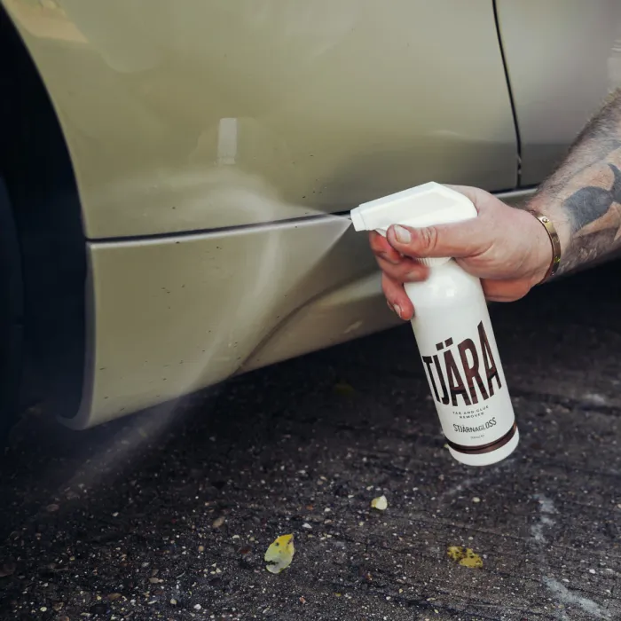 A hand sprays cleaning solution from a bottle labeled "TJÄRA" onto a car's lower side panel. The car is beige and situated on a gravel surface.