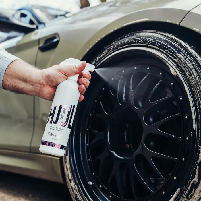 A hand sprays "HUL" color change wheel cleaner onto a wet, dirty black car wheel decorated with suds. The car is situated outdoors.