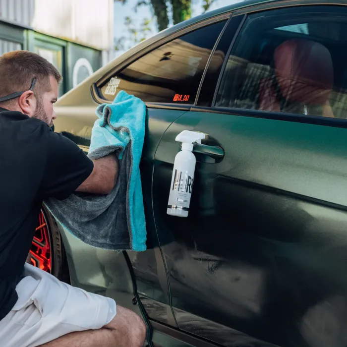 A person kneels, using a cloth to clean a metallic green car door, with a spray bottle of M1R Dorre Juice attached to the door handle in a parking area.