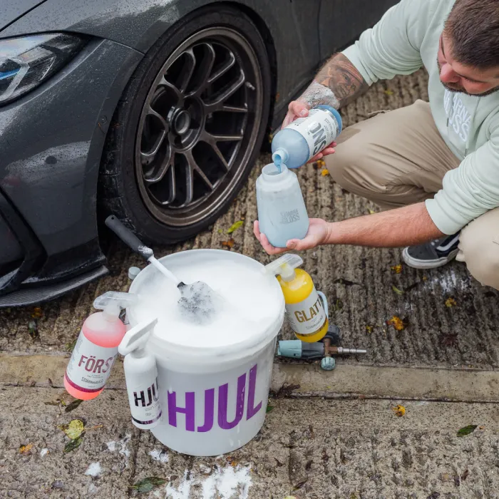 A person pours cleaning solution into a foaming pump next to a black car's wheel on a textured, wet ground. Nearby is a bucket with "HJUL" and bottles labeled "FÖRS" and "GLATT."