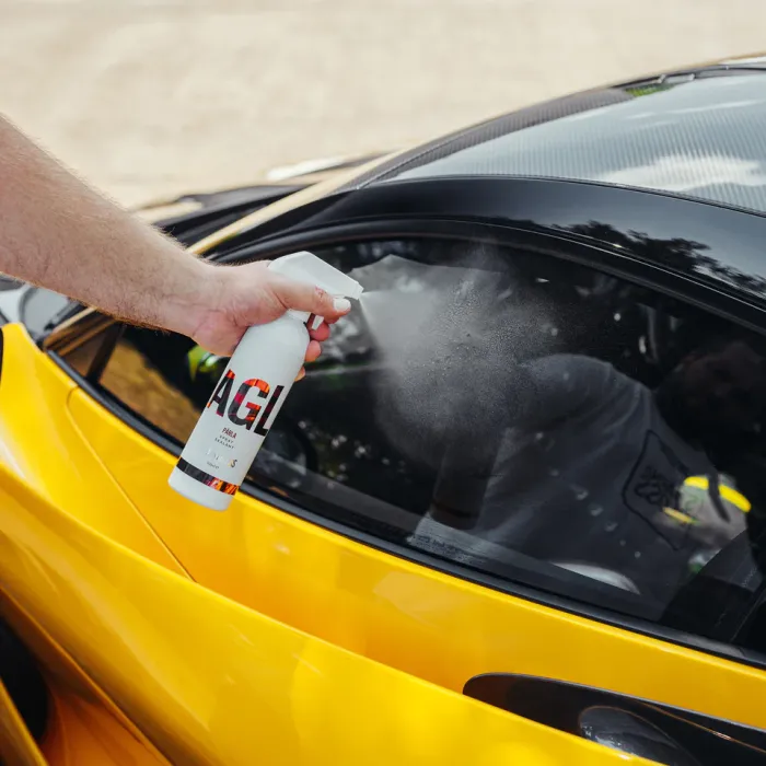 Hand sprays 'AGL' cleaner onto the black window of a yellow sports car, outdoors on a sunny day. Text on bottle: "AGL, RIVAL, Waterless Wash + Wax."
