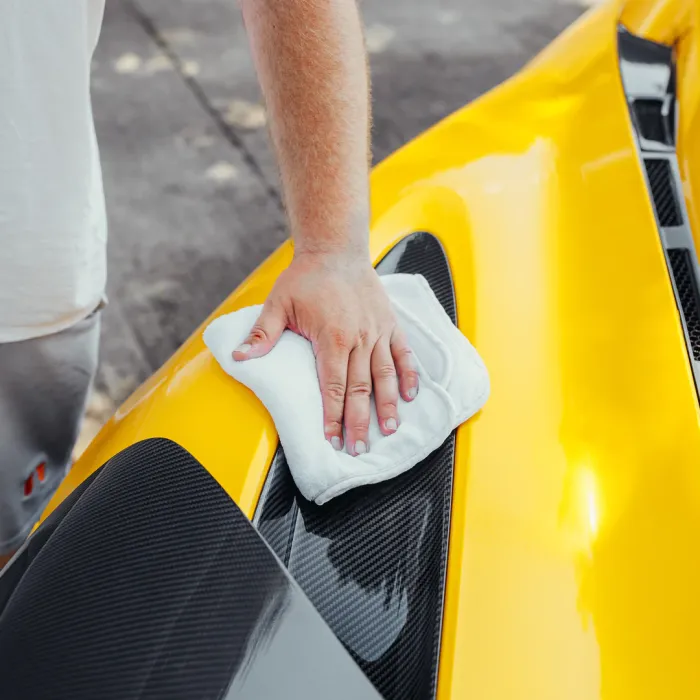 A hand is polishing a clean, shiny yellow car with a white cloth in an outdoor area, likely a driveway or parking lot.