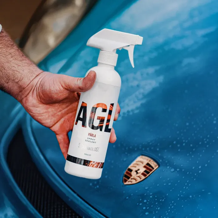 A hand holds a spray bottle labeled "AGL PÄRLA SPRAY SEALANT STARGLOSS" near the hood of a blue Porsche car.
