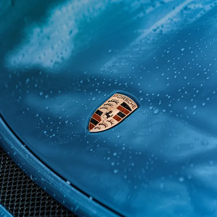 A close-up of a blue car's hood, with water droplets and the Porsche emblem prominently displayed at the center. Behind the hood, a black mesh grille is partially visible.