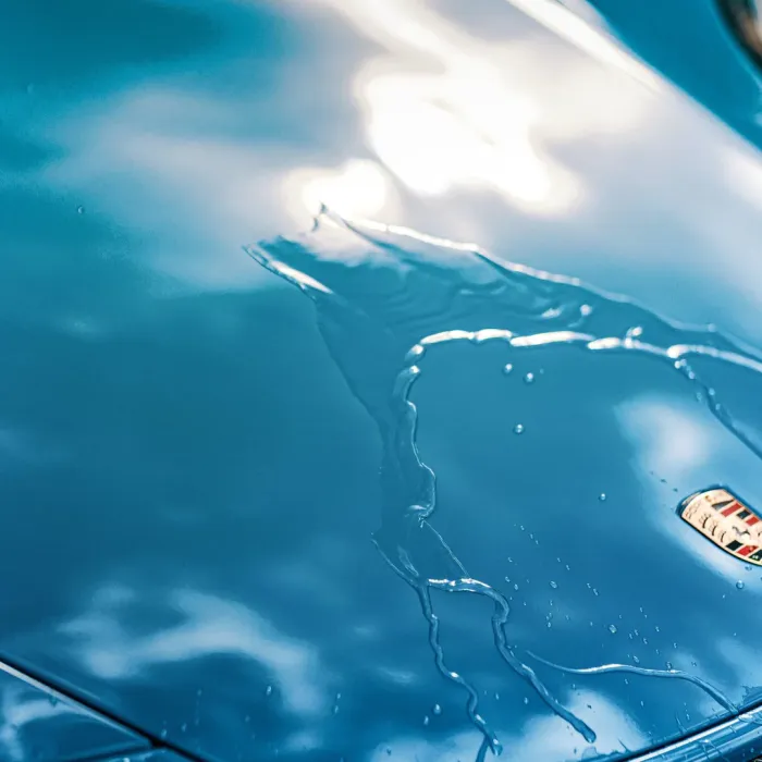 A blue car hood with water droplets forming a pattern, reflecting the sky and clouds, featuring a partial view of the car manufacturer’s logo.