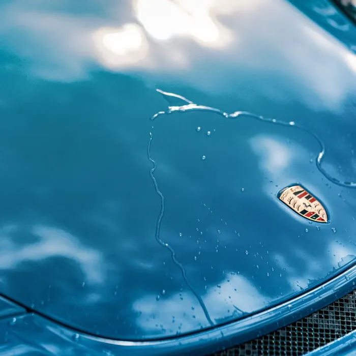 A water droplet glides down the shiny blue hood of a car, which features a Porsche emblem. The reflection of clouds can be seen on the hood’s surface.