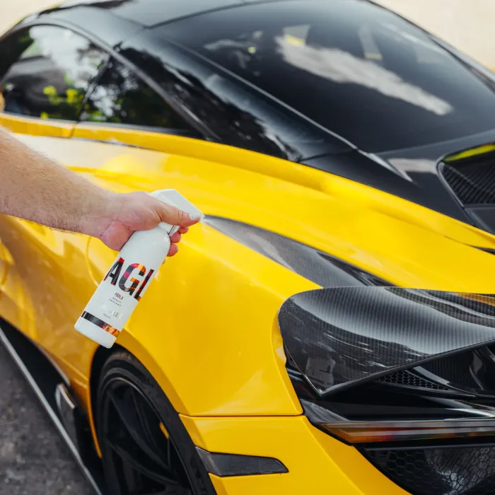 A hand sprays AGI Real liquid onto a yellow sports car's sleek exterior, under bright daylight reflecting off the car's shiny black panels.