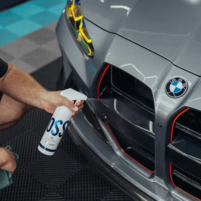 A hand sprays "OSS" cleaner onto the front grille of a grey BMW car in a well-lit, indoor setting with a textured floor.