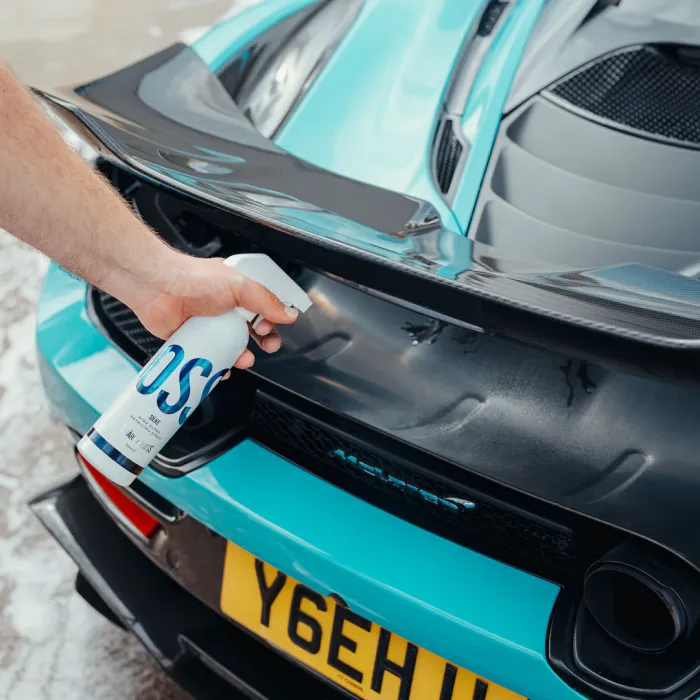 A hand is spraying cleaner from a bottle labeled "OSS" onto a blue sports car's rear end, with a yellow license plate reading "Y6EH." The car is parked on a concrete surface.
