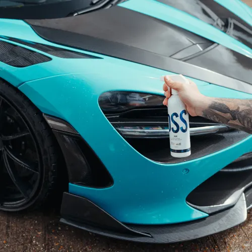 A hand sprays a product from a white can labeled "SOSS" onto a sleek, turquoise sports car's front bumper in an outdoor setting.