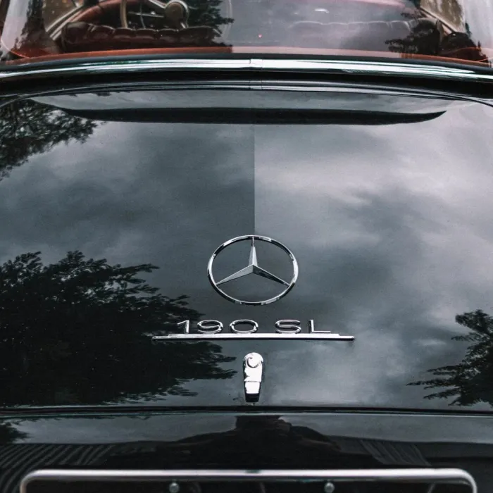 A black vintage car with a Mercedes-Benz emblem and "190 SL" text on the trunk, reflecting trees and a cloudy sky, with a partial view of the car's interior.