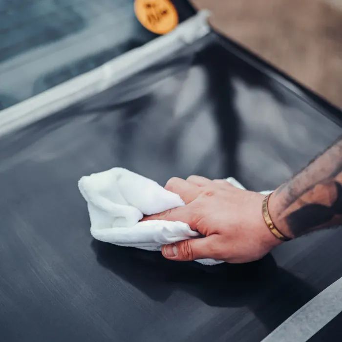 A hand holding a white cloth is wiping a black surface, likely a car window, in an outdoor setting with a yellow sticker visible on the vehicle.