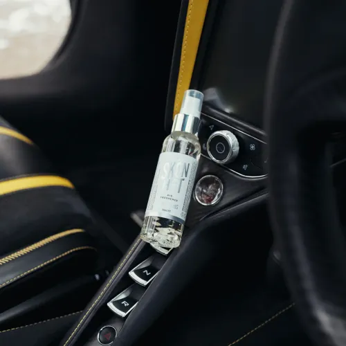 A bottle of SKON HET air freshener is positioned upright, leaning against the central control console inside a car with black and yellow interior.