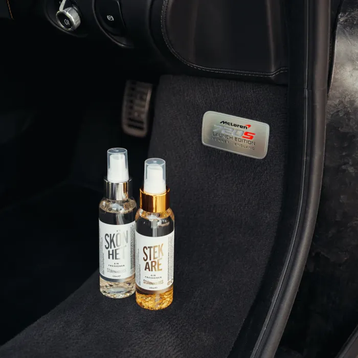 Two spray bottles labeled "SKÖNHET" and "STEKARE" standing in a car's footwell with pedals; a metal badge reads "McLaren 675LT Launch Edition, Woking, England."