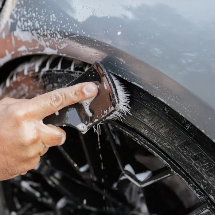 A hand uses a soapy brush to scrub the soapy surface of a car tire and fender, which is covered in dark, wet mud from a recent wash.