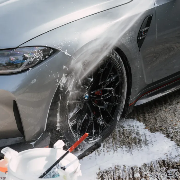 A silver car's front wheel is being sprayed with water and soap, next to a plastic bucket filled with cleaning tools, on a wet concrete surface.