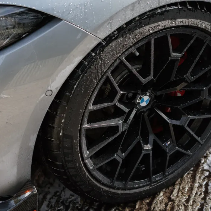 A wet black BMW wheel with intricate spoke design, displaying the brand logo, is attached to a silver car body, situated on a wet, textured surface, tire labeled "MICHELIN Pilot Sport 4S".