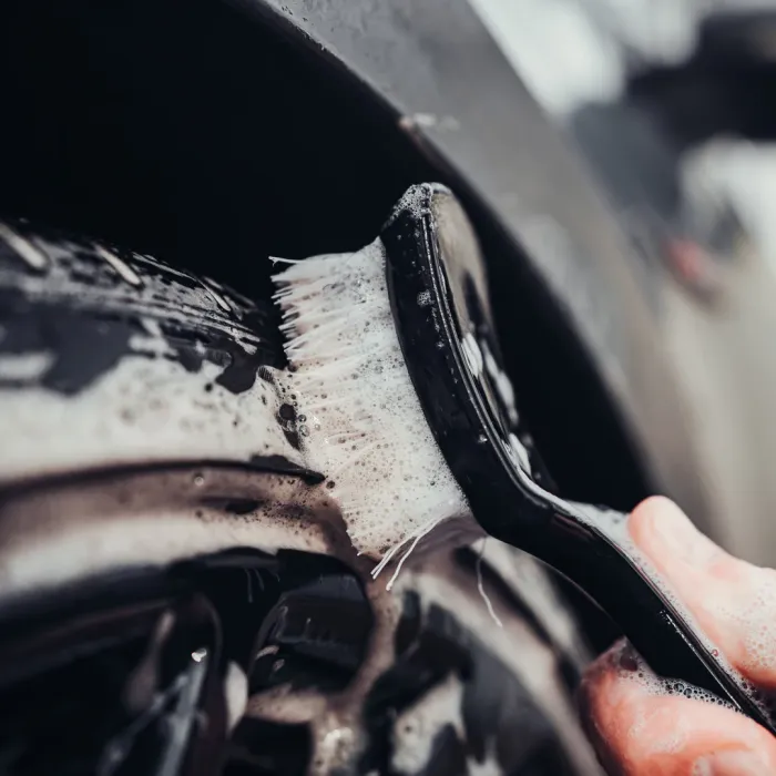 A hand uses a soapy brush to clean a car tire, focusing on removing dirt and grime, with the car's exterior visible in the background.