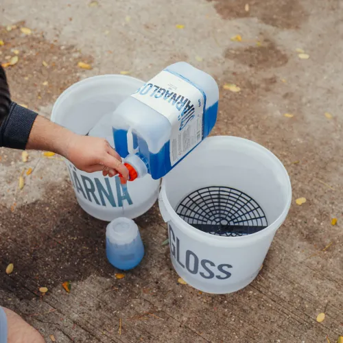 A person pours blue liquid from a container labeled "MARANGA" into a smaller bottle, beside two buckets labeled "ARNA" and "GLOSS," on a concrete surface scattered with small leaves.