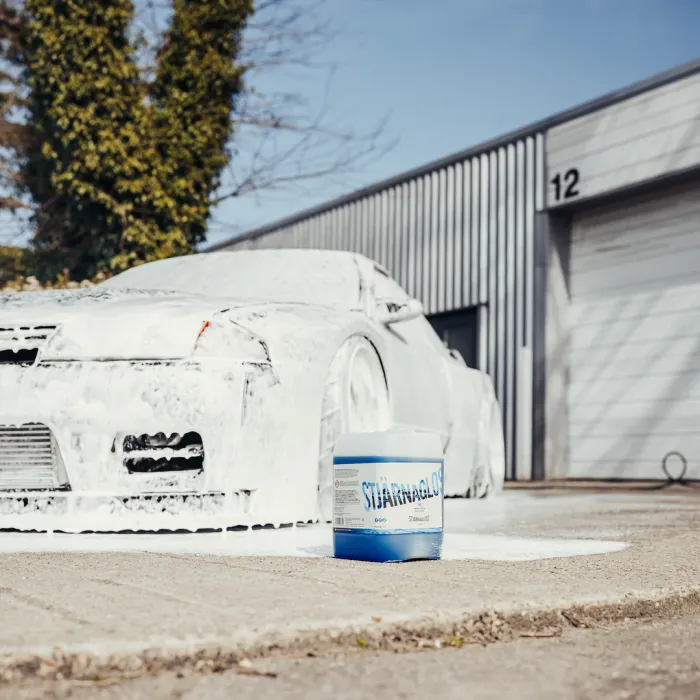 A white car covered in foam stands near a grey industrial building with a closed garage door marked "12." In the foreground, a blue and white container labeled "STJÄRNGLANS."