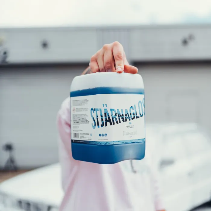 A hand holds a container labeled "STJÄRNAGLOSS" with blue liquid, positioned against a blurry background of an outdoor industrial area.